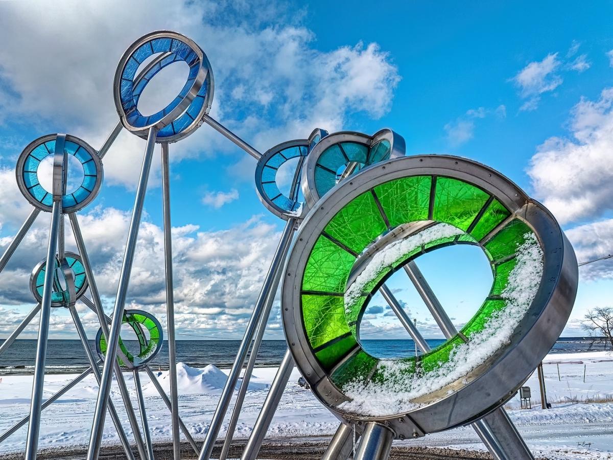 Public art installation featuring circular metal structures with green glass accents, located by a snowy lakeside under a partly cloudy sky.