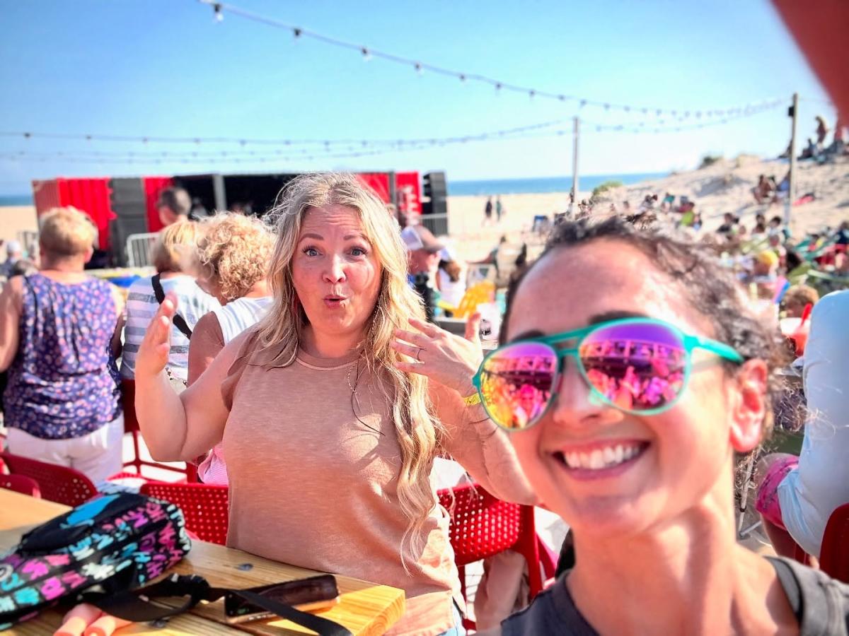 Two people sitting at a table at The Deck, smiling and making playful expressions at an outdoor beach setting with others in the background and string lights visible overhead.