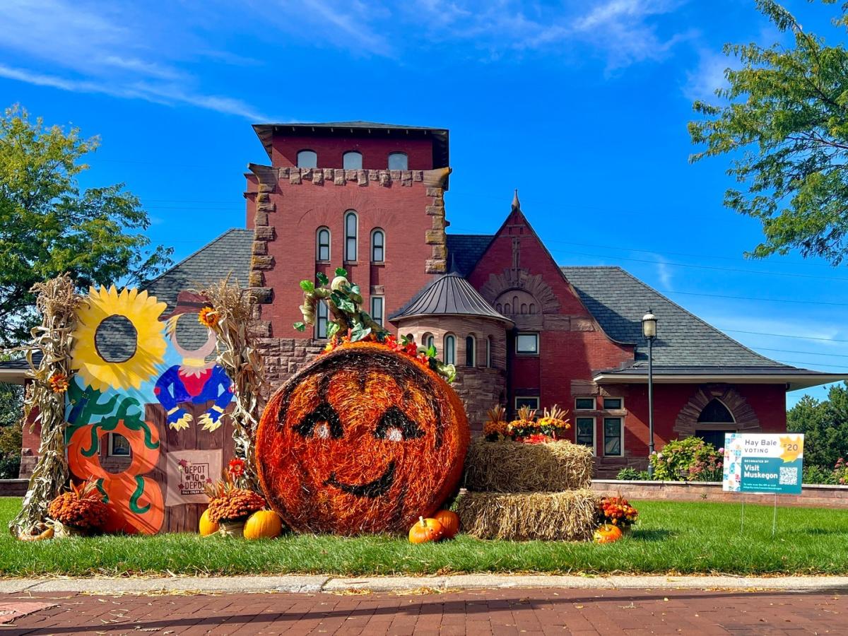 An autumn display in front of a historic red brick building, Union Depot in downtown Muskegon, featuring a large hay bale painted to resemble a smiling pumpkin, adorned with sunflowers, colorful autumn leaves, and smaller pumpkins arranged around. A sign reads Vote for Me