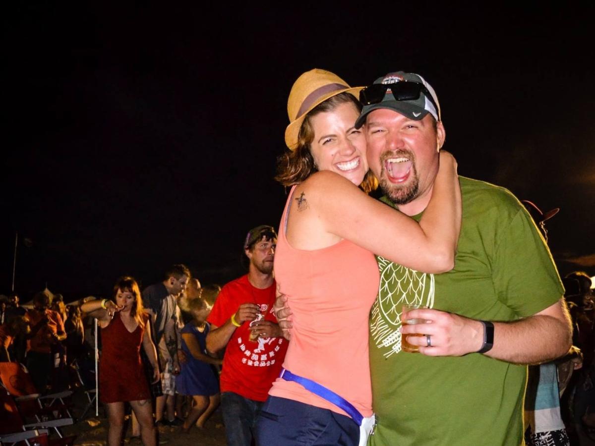 Two people joyfully embracing at an outdoor night event, burning foot beach festival, one holding a beverage, with others in the background enjoying the festivities.