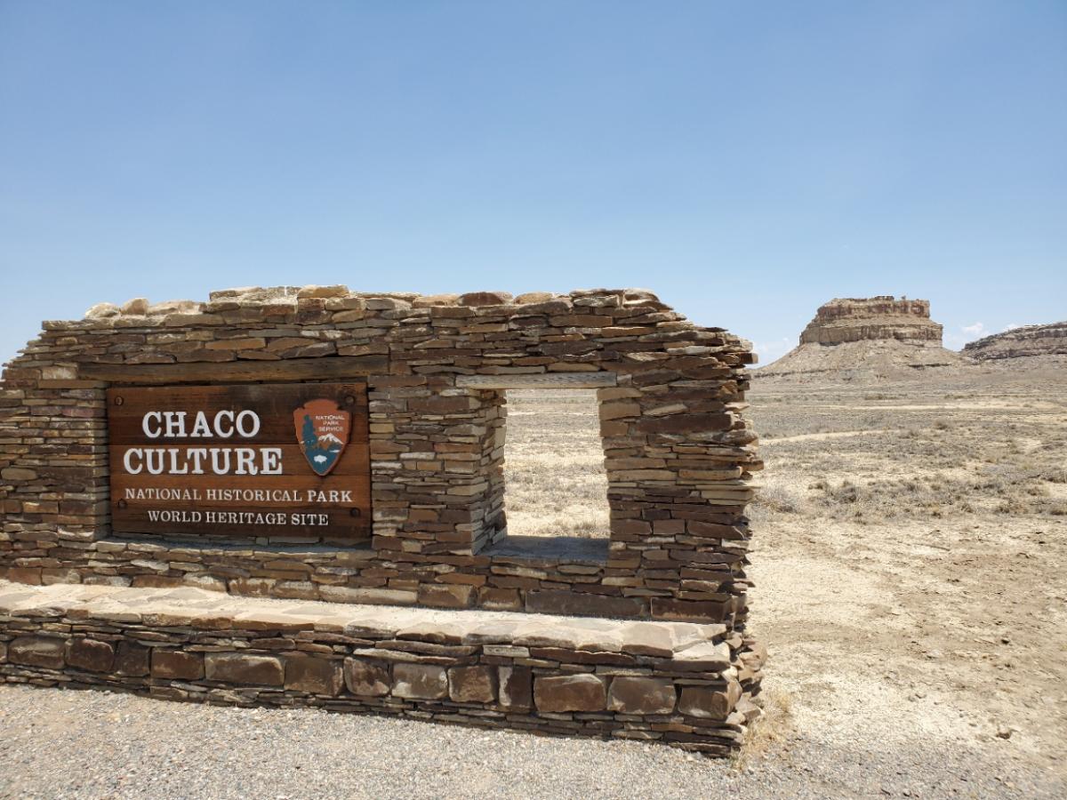 Sign reading "Chaco Culture National Historical Park, World Heritage Site" mounted on a low stone wall, with a clear blue sky and a flat-topped mesa in the background.
