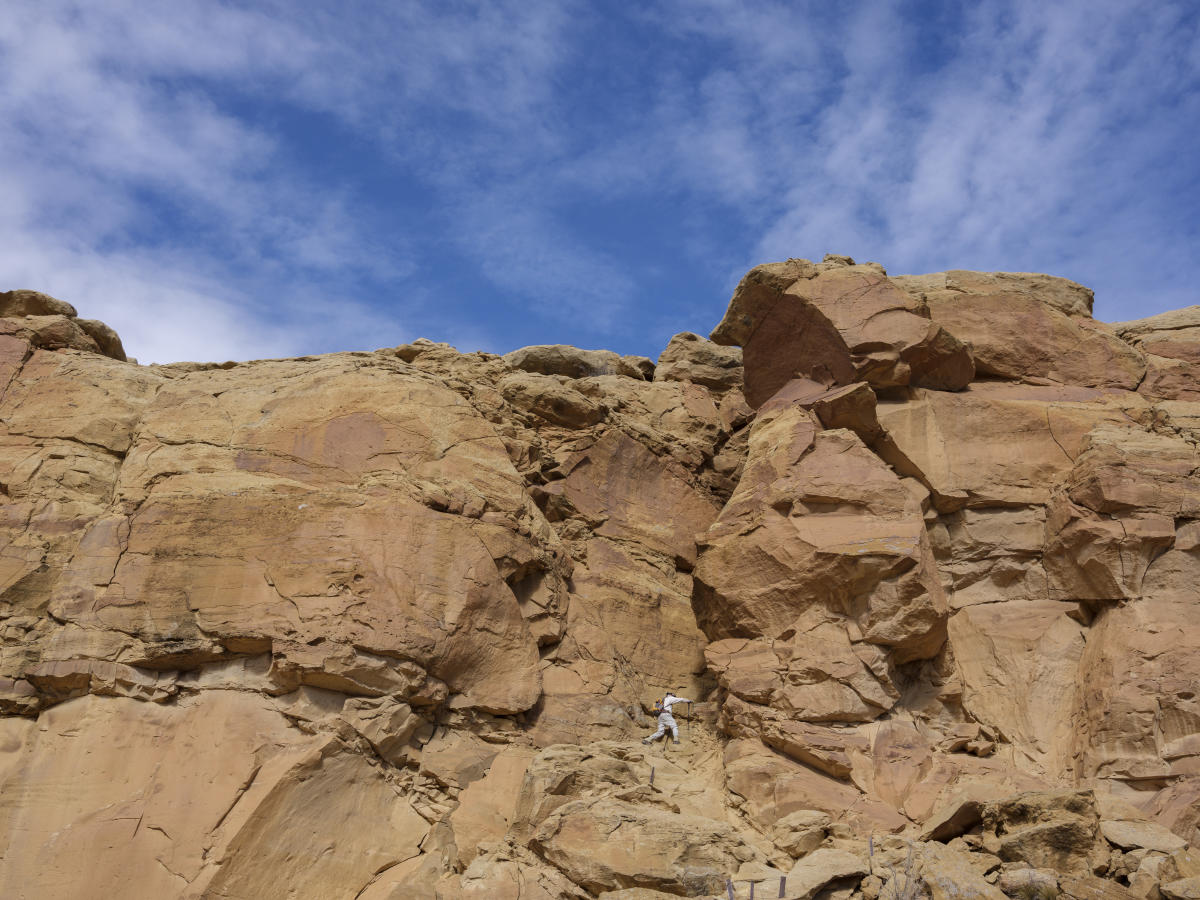 Climber ascends a large, rugged sandstone cliff under a vibrant blue sky.