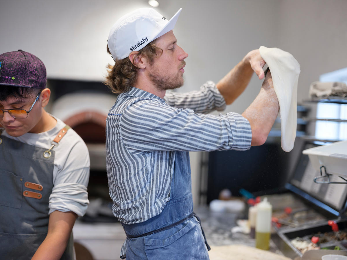 A person in a striped shirt and baseball cap stretches pizza dough in a bright kitchen, while another person in an apron prepares ingredients nearby.