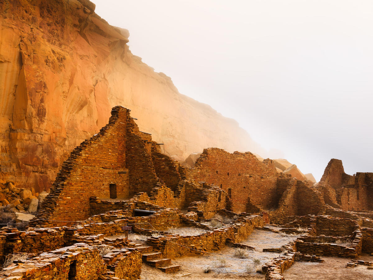 The remains of stone buildings, part of ancient ruins, stand in front of a tall, sheer cliff face.