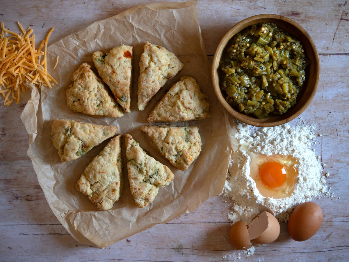 A flatlay of ingredients and baked goods on a wooden surface. There is a bowl of chopped green peppers, an egg in a pile of flour, a bowl of shredded cheese, and eight baked scone triangles on parchment paper.