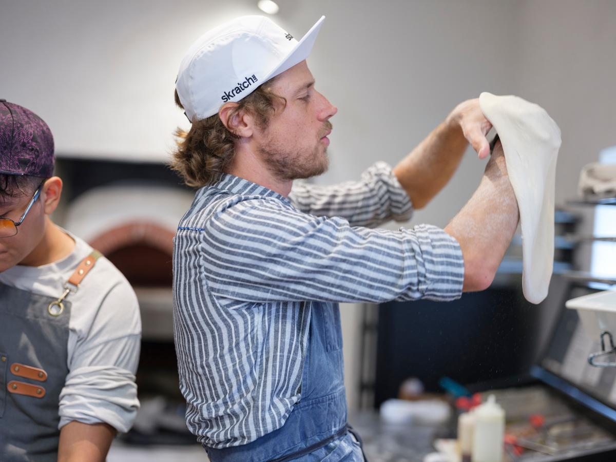 A person in a striped shirt and baseball cap stretches pizza dough in a bright kitchen, while another person in an apron prepares ingredients nearby.
