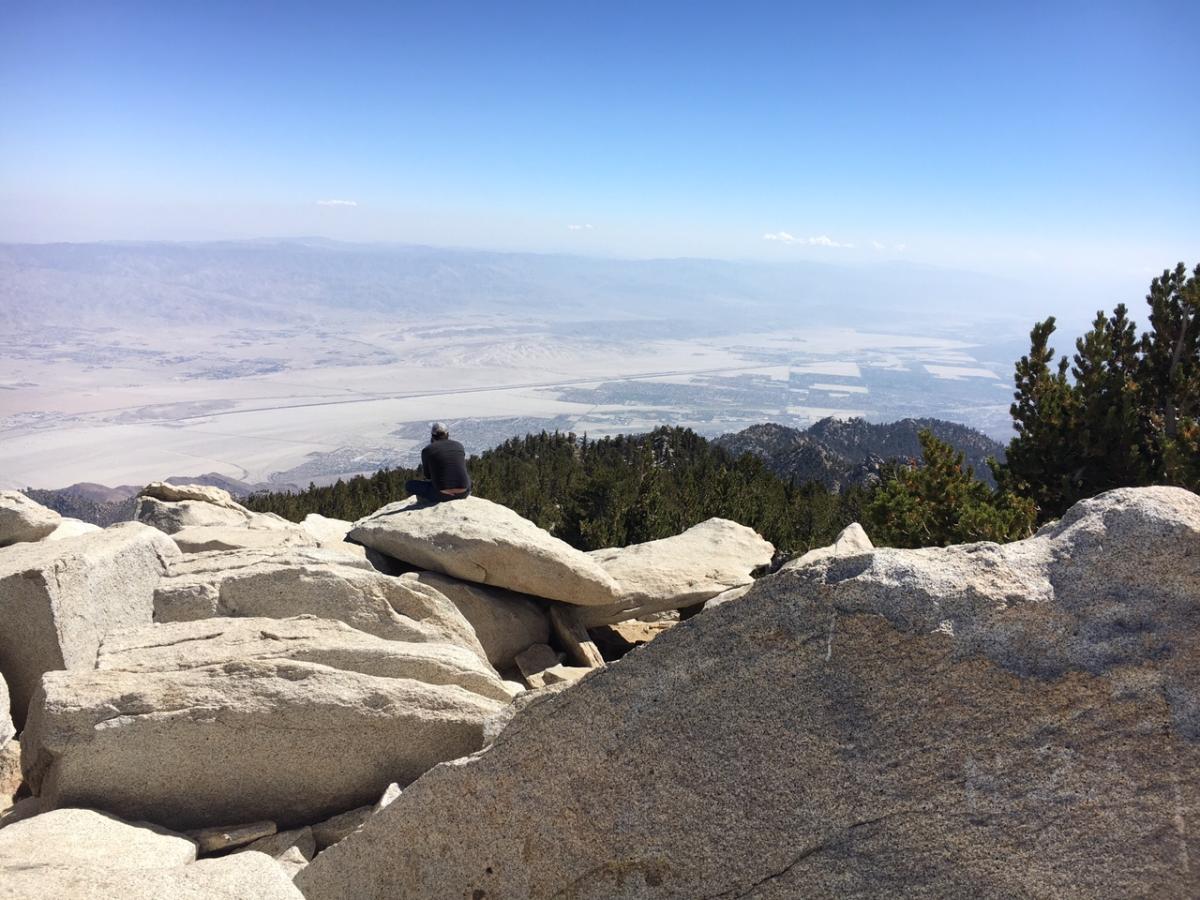 A hiker sits on a boulder on San Jacinto Peak overlooking the desert below