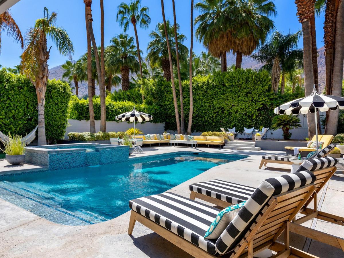 A stylish outdoor space with striped lounge chairs, a sparkling blue swimming pool, umbrellas, and palm trees in a sunny setting.