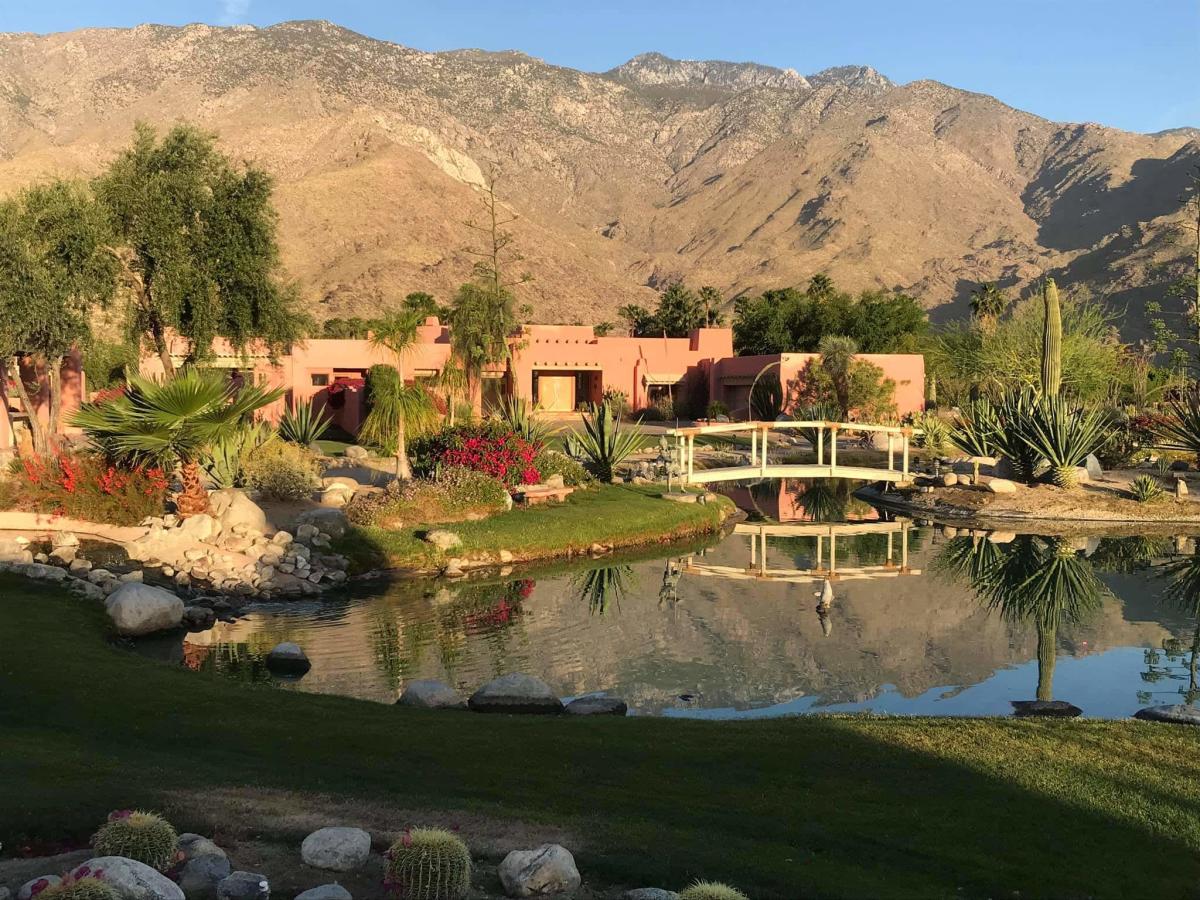 A serene landscape featuring lush gardens, a reflective pond with a white footbridge, terracotta-colored buildings, and a backdrop of rugged mountains under a clear sky.