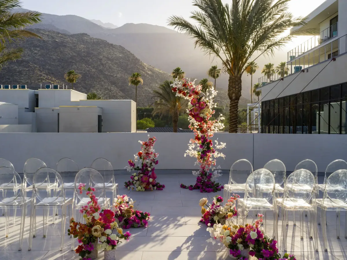 Outdoor wedding ceremony setup with clear ghost chairs and flower arrangements on a sunny day, mountains in the background, and palm trees adjacent to a modern building.