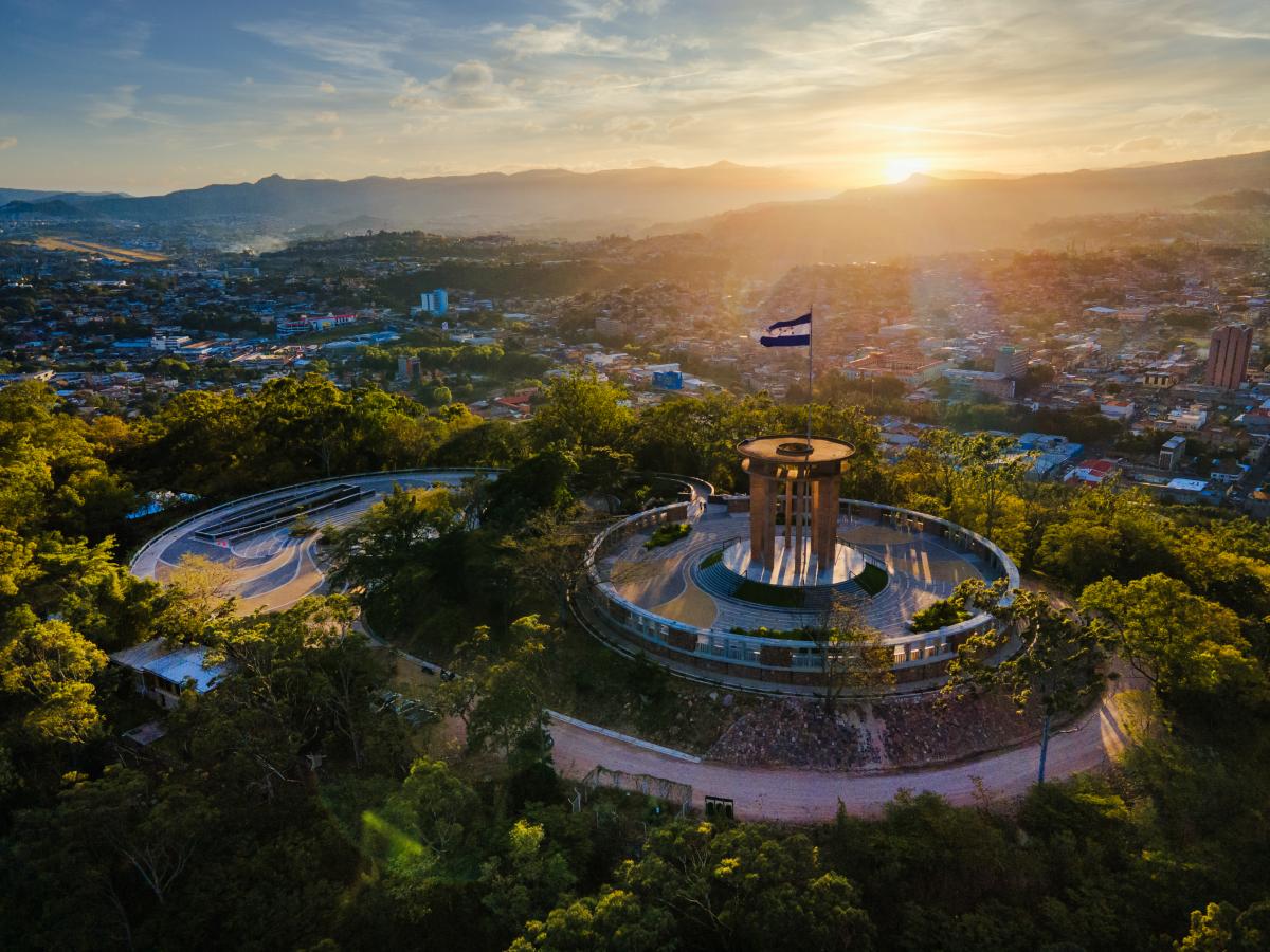 An aerial view of a circular monument with a flagpole at its center. The monument is  surrounded by trees and overlooks a city at sunset. The sky is filled with colorful clouds