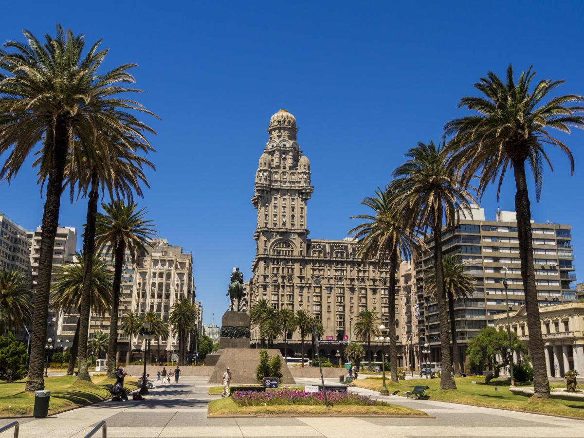 Alt Text: A grand, ornate building with a dome and a tower stands tall in a city square.  Palm trees line the sides of the square, and a statue of a man on horseback is visible in the  foreground.