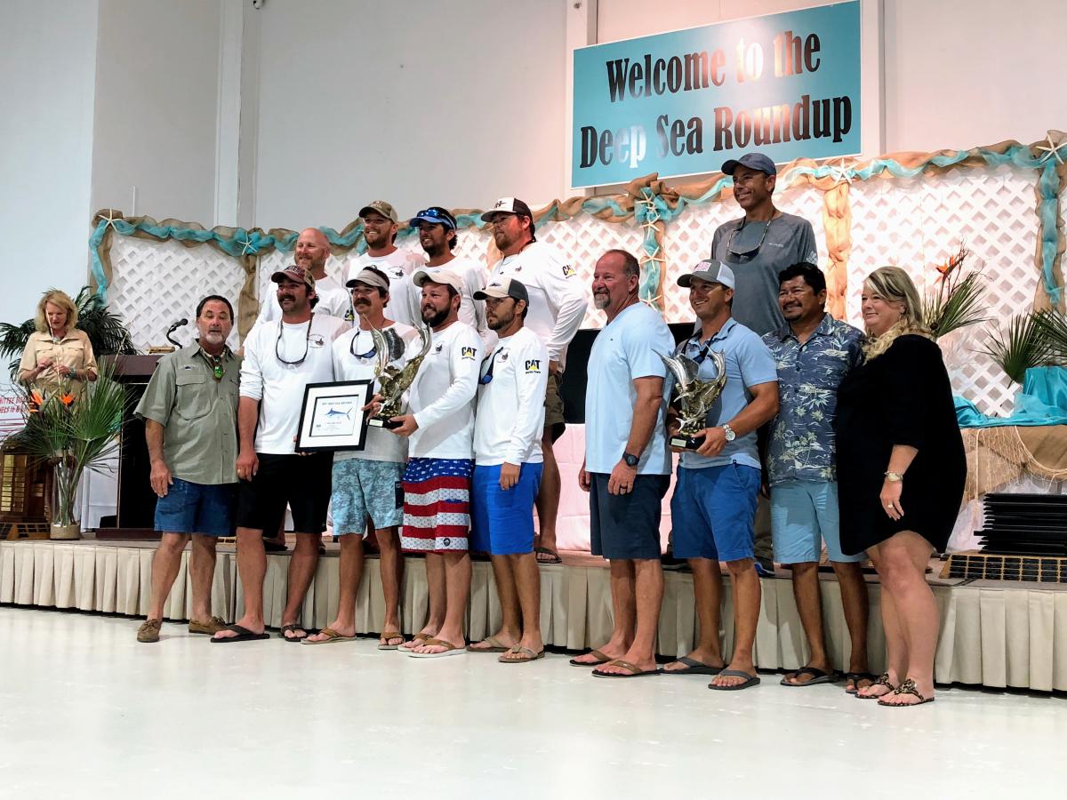 A group of anglers pose for a photo in front of a stage, holding a trophy and certificate