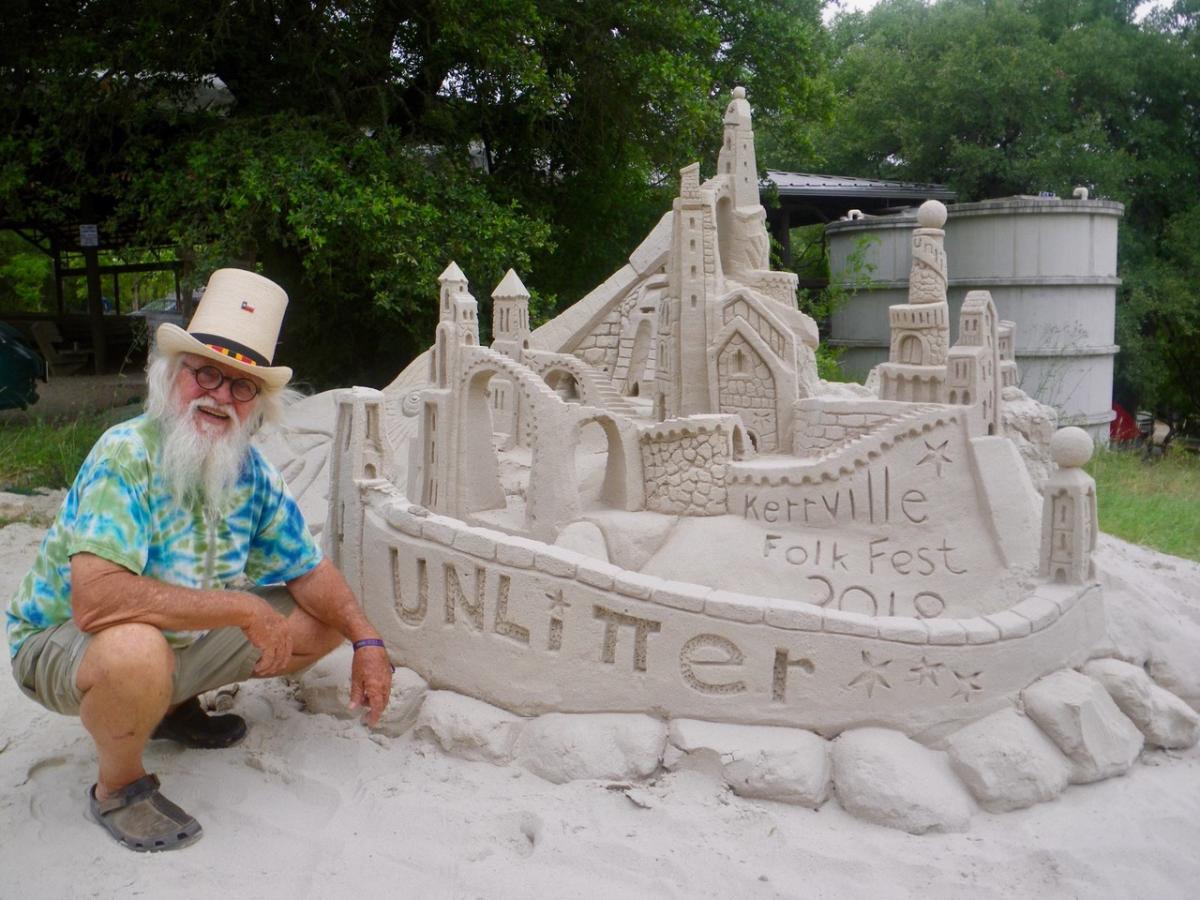 An old man in a straw tophat squats next to a large sandcastle. The front of the casgtle says, "Unlitter."