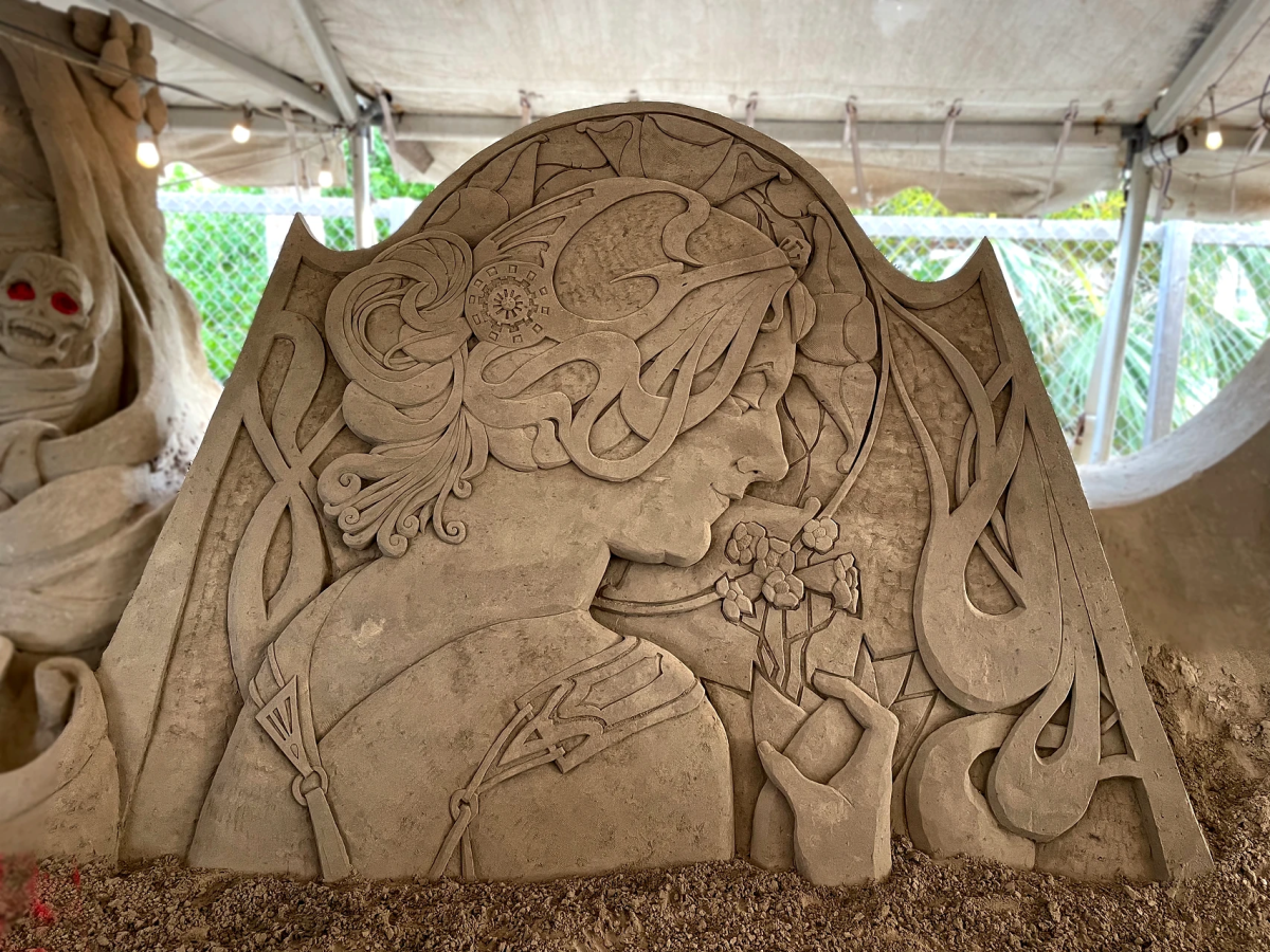 Sand sculpture of a woman holding a bundle of flowers and sniffing them surrounded by delicate designs.
