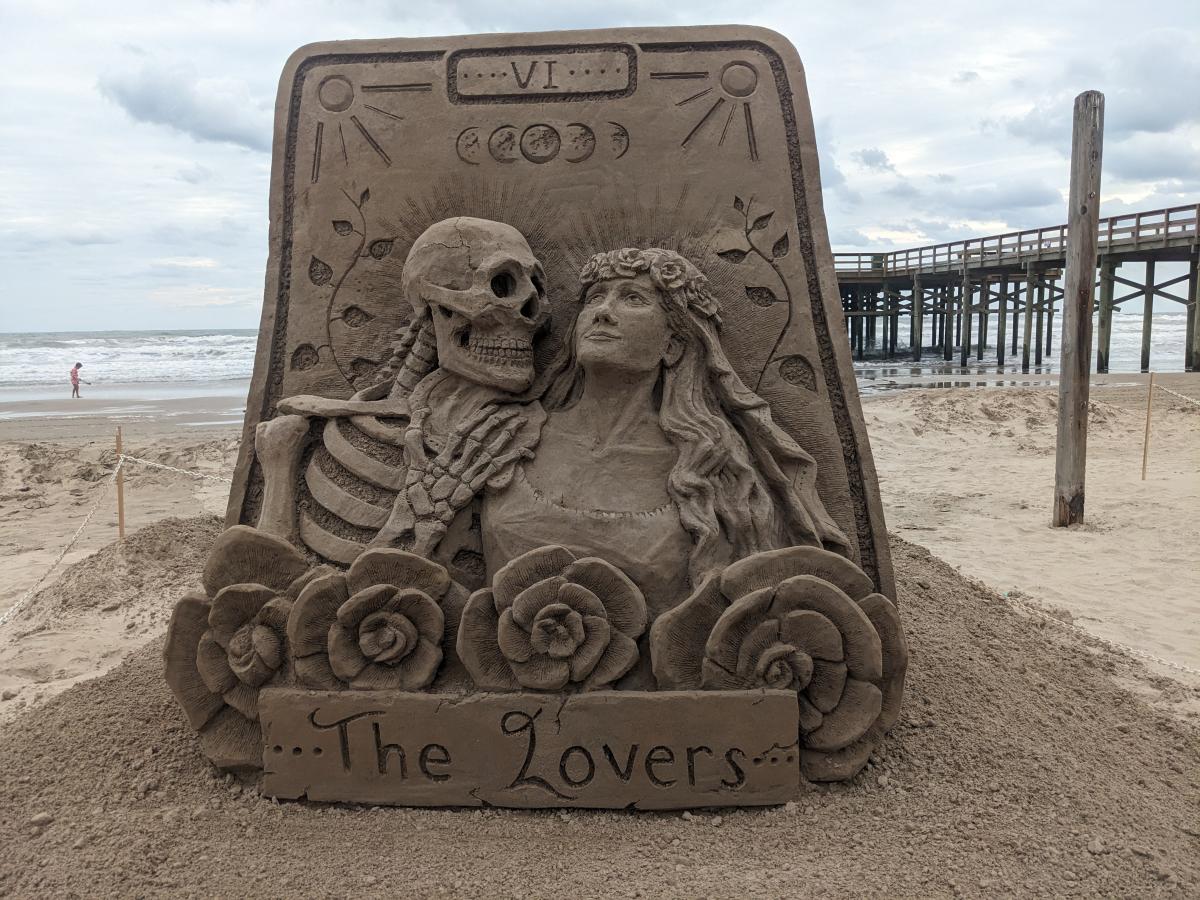 Sand sculpture in the shape of a card that has two figures coming out of it—a skeleton and a woman. Underneath is a banner reading, "The Lovers."