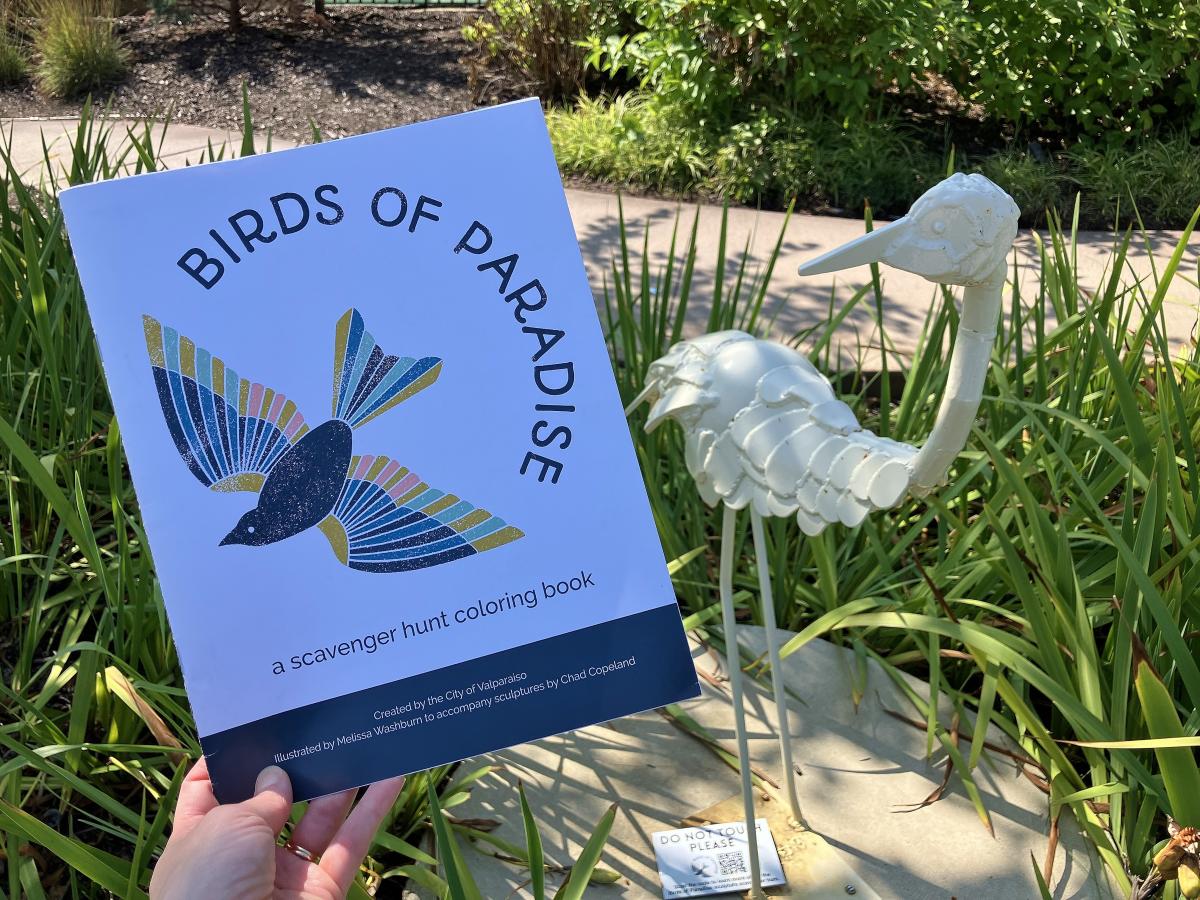 A hand holds a brochure with an illustrated bird and "Birds of Paradise" nest to a white bird sculpture.