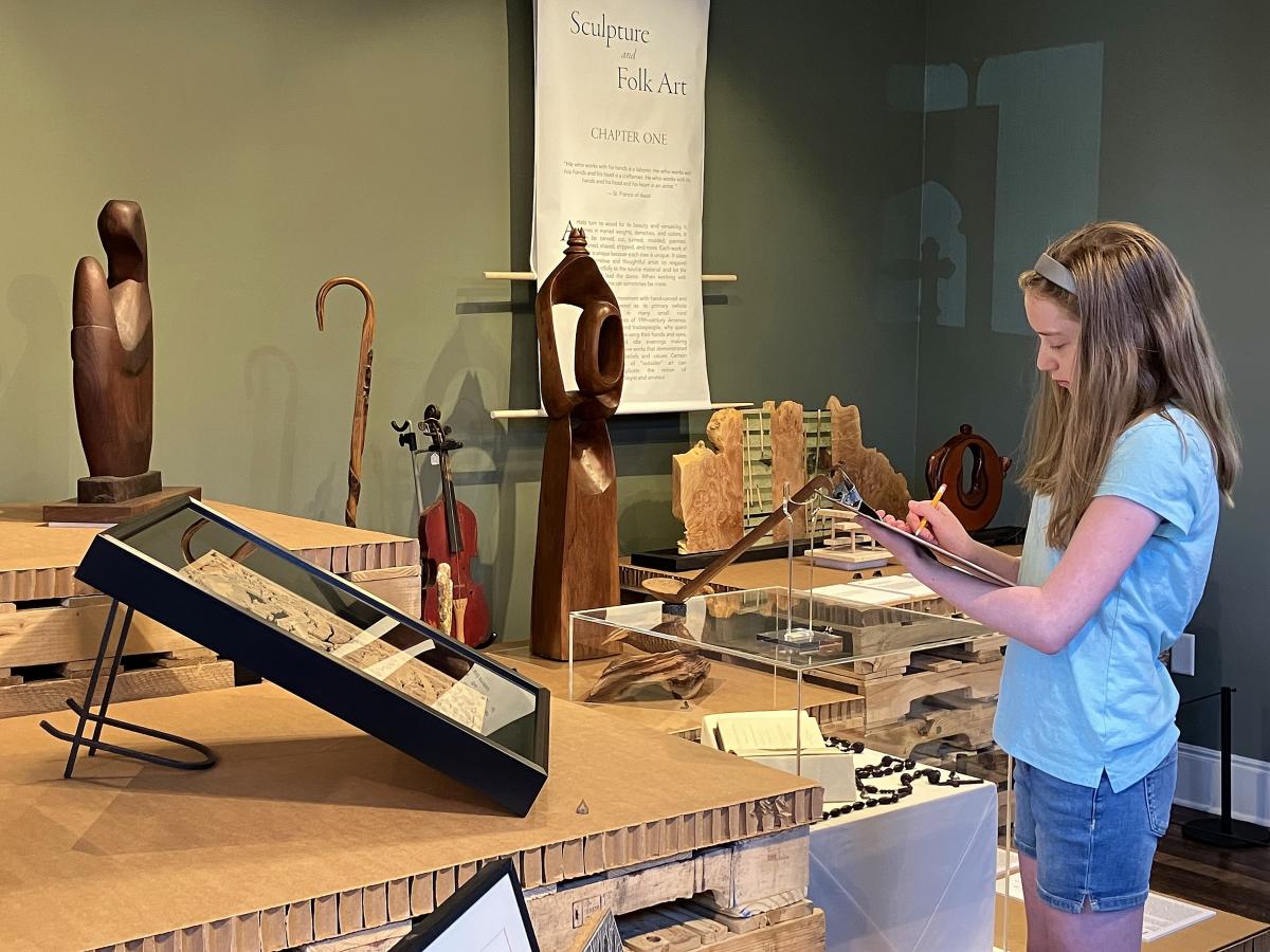 A young woman stands in front of a museum exhibit, taking notes. She is wearing a light blue shirt and jean shorts.