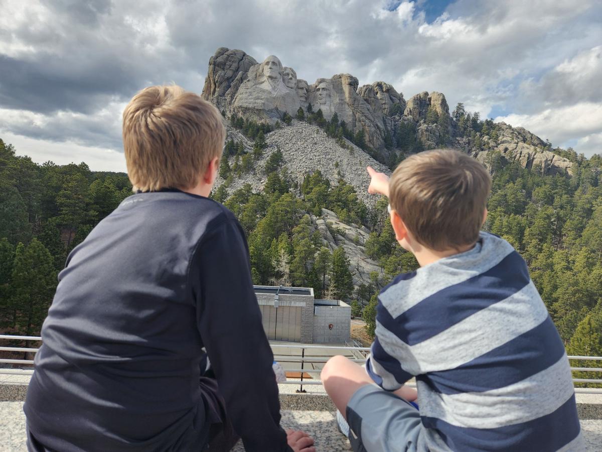 two kids sitting on the grand view terrace at mount rushmore pointing at the memorial