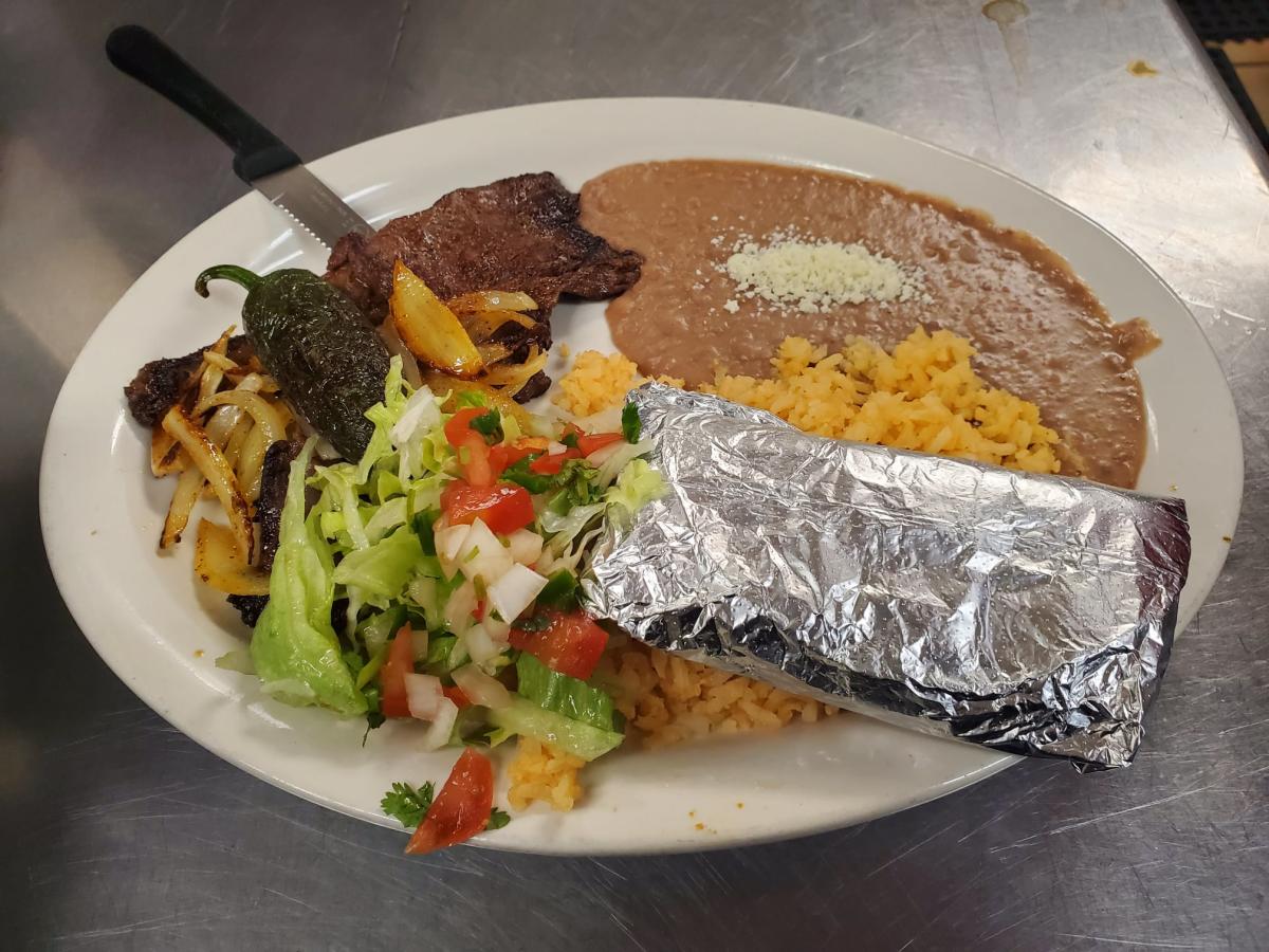 A large white oval plate on a stainless steel counter holds a Mexican-style meal with a foil-wrapped burrito, Mexican rice, refried beans topped with crumbled white cheese, grilled steak with sautéed onions, a whole roasted jalapeño pepper, and a small side salad of lettuce, tomato, and chopped onion. A steak knife rests on the edge of the plate.