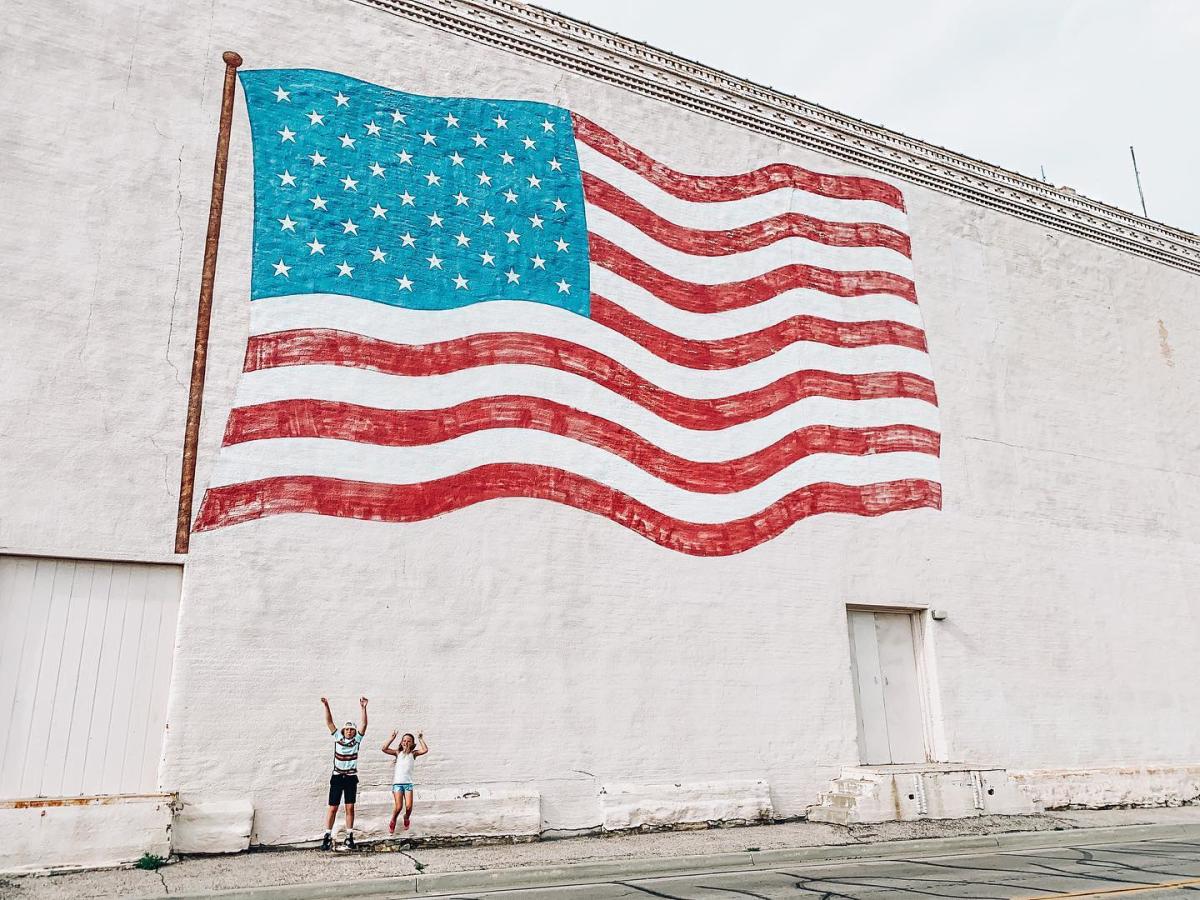 A large mural of the American flag painted on a white wall. Two children stand joyfully with arms raised beneath it, conveying a patriotic and cheerful mood.