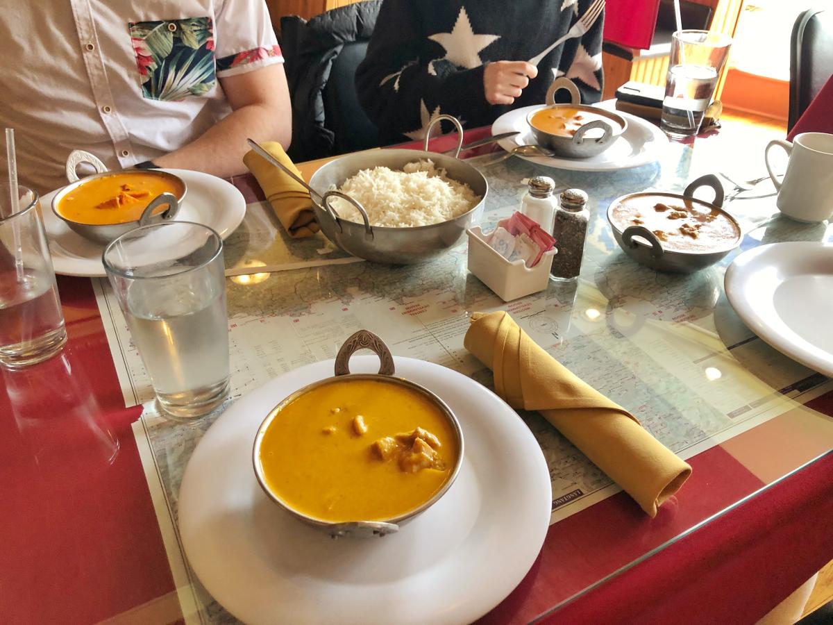 A table with Indian curry dishes and white rice in metal bowls, surrounded by glasses of water. Diners wear casual clothing, creating a cozy atmosphere.