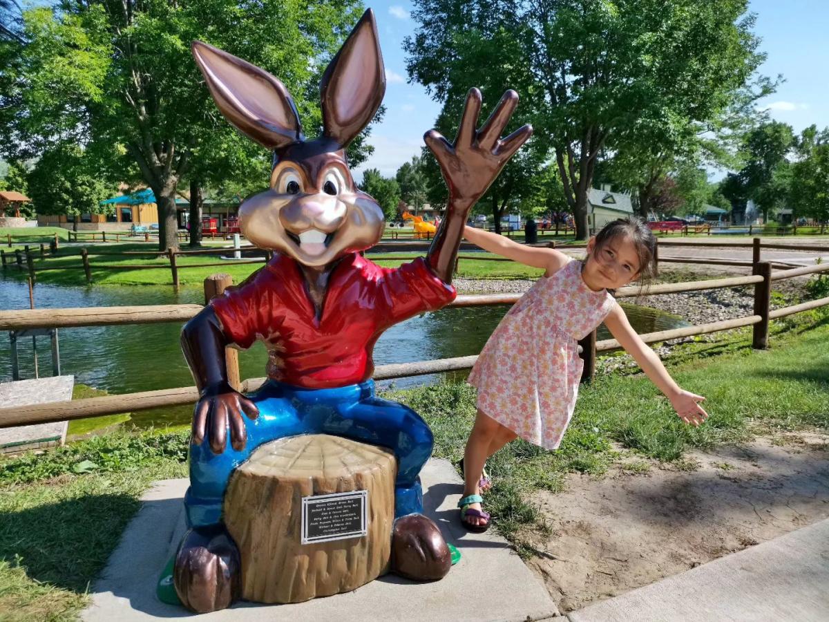 A smiling girl in a floral dress poses playfully beside a large colorful rabbit statue, set in a sunny park with lush trees and a pond.