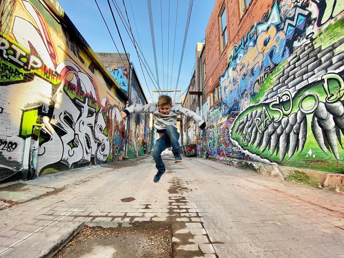 A child joyfully jumps mid-air in the vibrant Art Alley filled with colorful graffiti art on brick walls. Power lines stretch across the blue sky above.