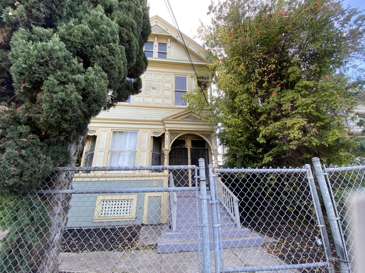 A yellow Victorian-era home is preserved by a fence and surrounded by lush green trees