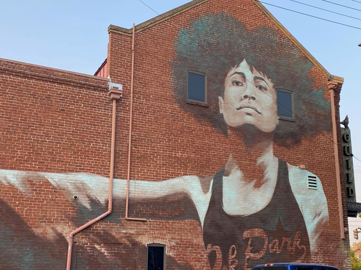 A black-and-white mural of a woman with a tank top and afro textured hair adorns the side of a brick building in the neighborhood of Oak Park