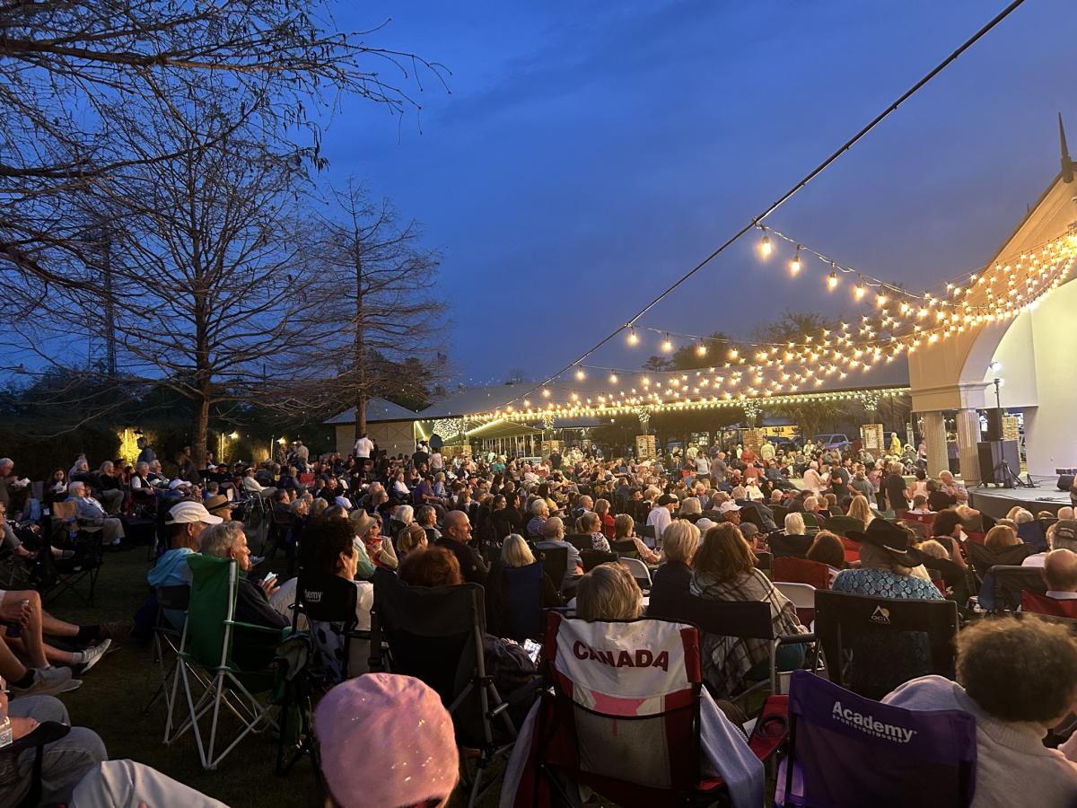 Large crowd seated in lawn chairs at Mandeville Trailhead during a Mandeville Live! Free Friday concert, with string lights glowing overhead and the amphitheater stage illuminated at dusk.