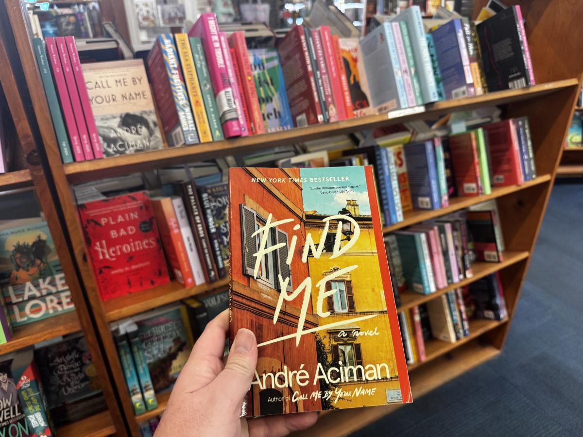 A hand showing of the cover of a novel in front of shelves lined with colorful books