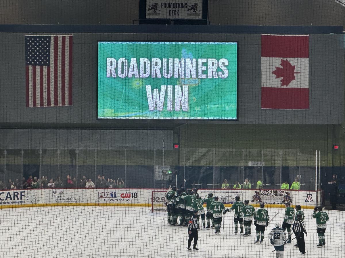 Hockey players on an ice rink in front of a scoreboard reading "ROADRUNNERS WIN," with American and Canadian flags displayed above.