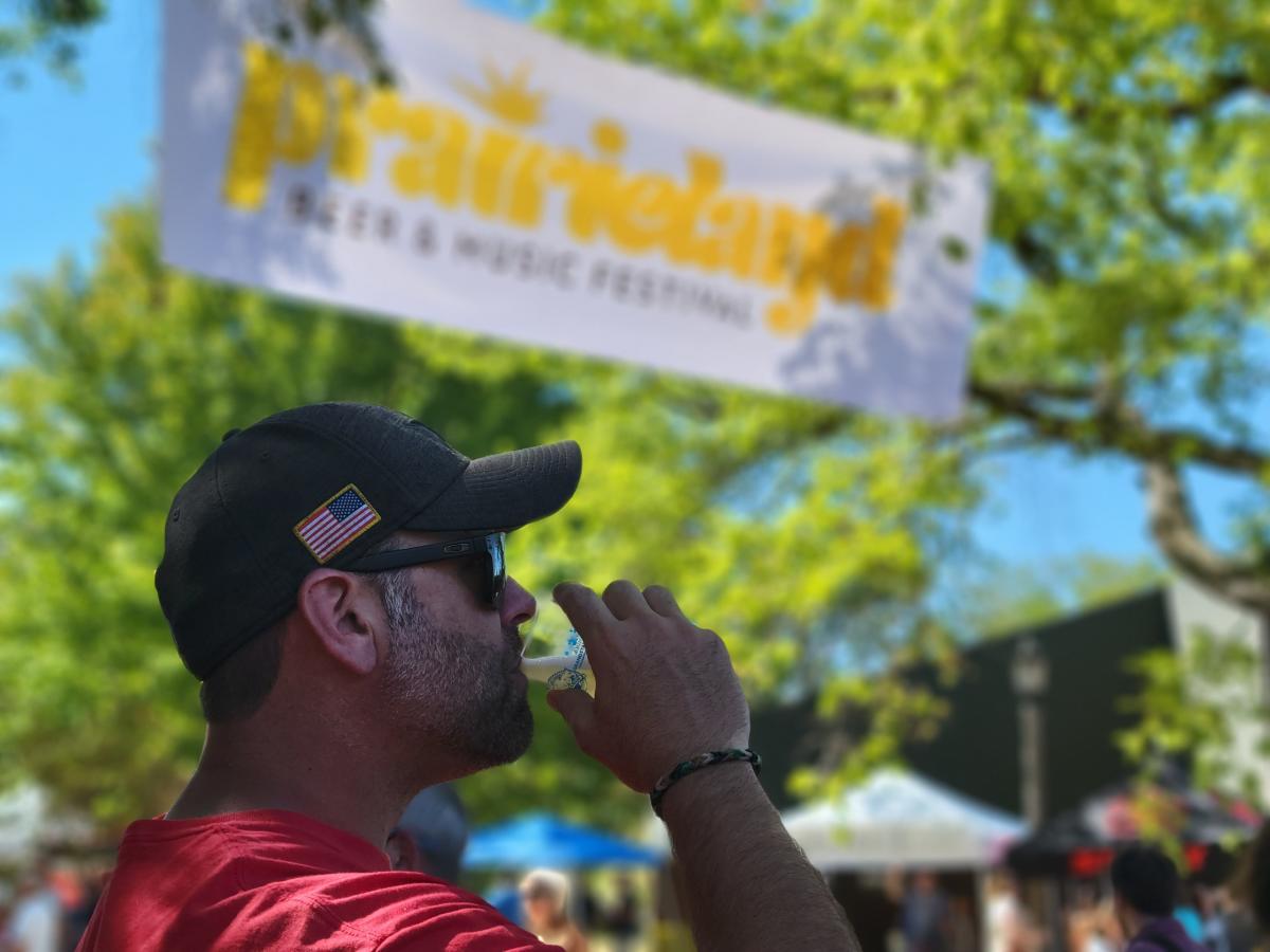A man drinks a beer under a banner for Prairieland Beer & Music Festival