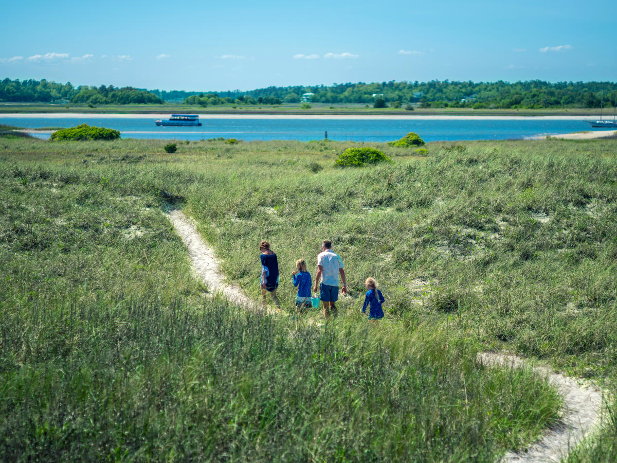 Family of four walking on a path on Masonboro Island with the Carolina Runner sailing in the background.
