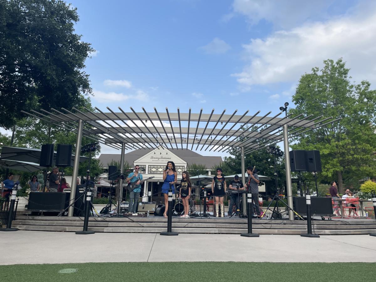 The camera faces the stage of Market Street's Central Park during National Travel and Tourism Week 2024. There is a band on the stage, but Elizabeth Eddins holds the microphone to address the crowd (who are off-screen).