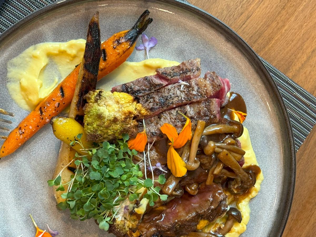 The camera focuses on an expertly plated meal at The Waterway Marriott. It features a nearly rare steak, cut into strips, and it's topped with a BBQ-like sauce, long and skinny mushrooms, orange flower petals, and sprouts. It is served with several cooked carrots (yellow and orange) and cooked cauliflower, and it's served on a thick yellow saffron sauce.