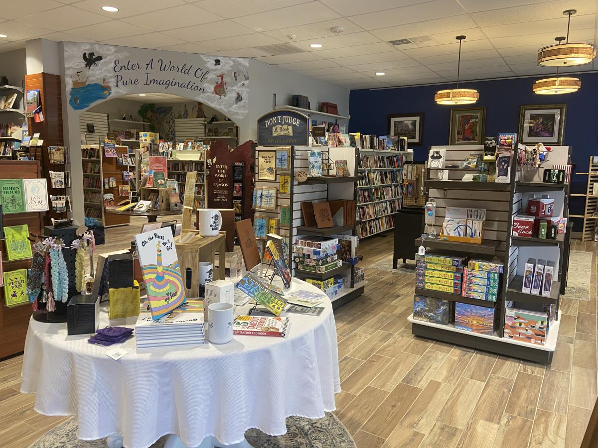 The camera offers a wide-angle look at Buy The Book. On the right, there are two square display stands with 2-3 shelves on each side, each featuring various puzzles, notebooks, and other knicknacks. Behind those are 2-3 long bookshelves cram-packed with books. On the wall behind those are several book-themed paintings in thick frames. On the left side of the photo, there is a large white-clothed table filed high with books, bookmarks, mugs, and hair accessories. Further back and to the left is a room full of books for younger readers. Above the arched doorway, the wall is painted with clouds and various fairytale creatures surrounding text that reads, "Enter a World of Pure Imagination."