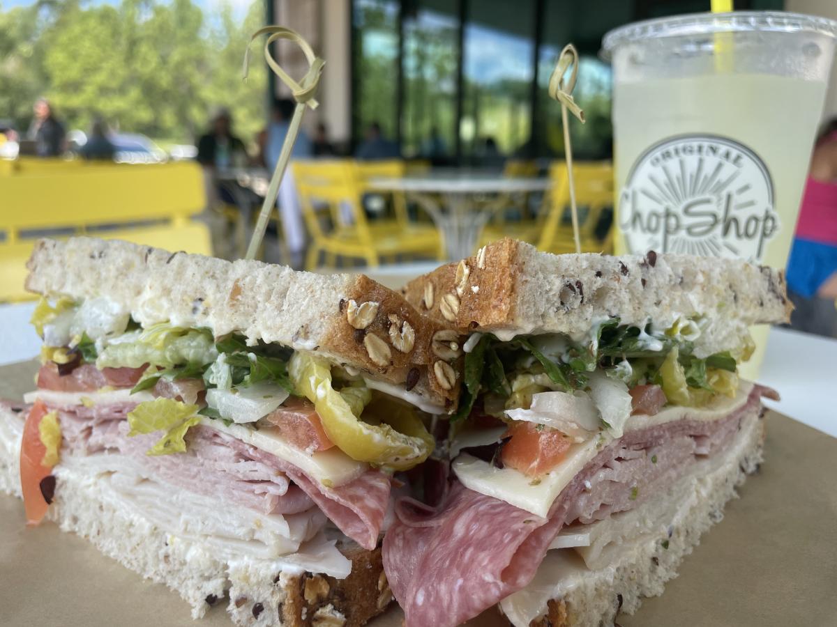 On the outdoor patio of Original ChopShop, two halves of a layered salami, swiss, and bell-pepper sandwich face the outer edges of the camera frame. Ice-cold lemonade in an Original Chop-Shop to-go cup is slightly out of focus behind the sandwich.