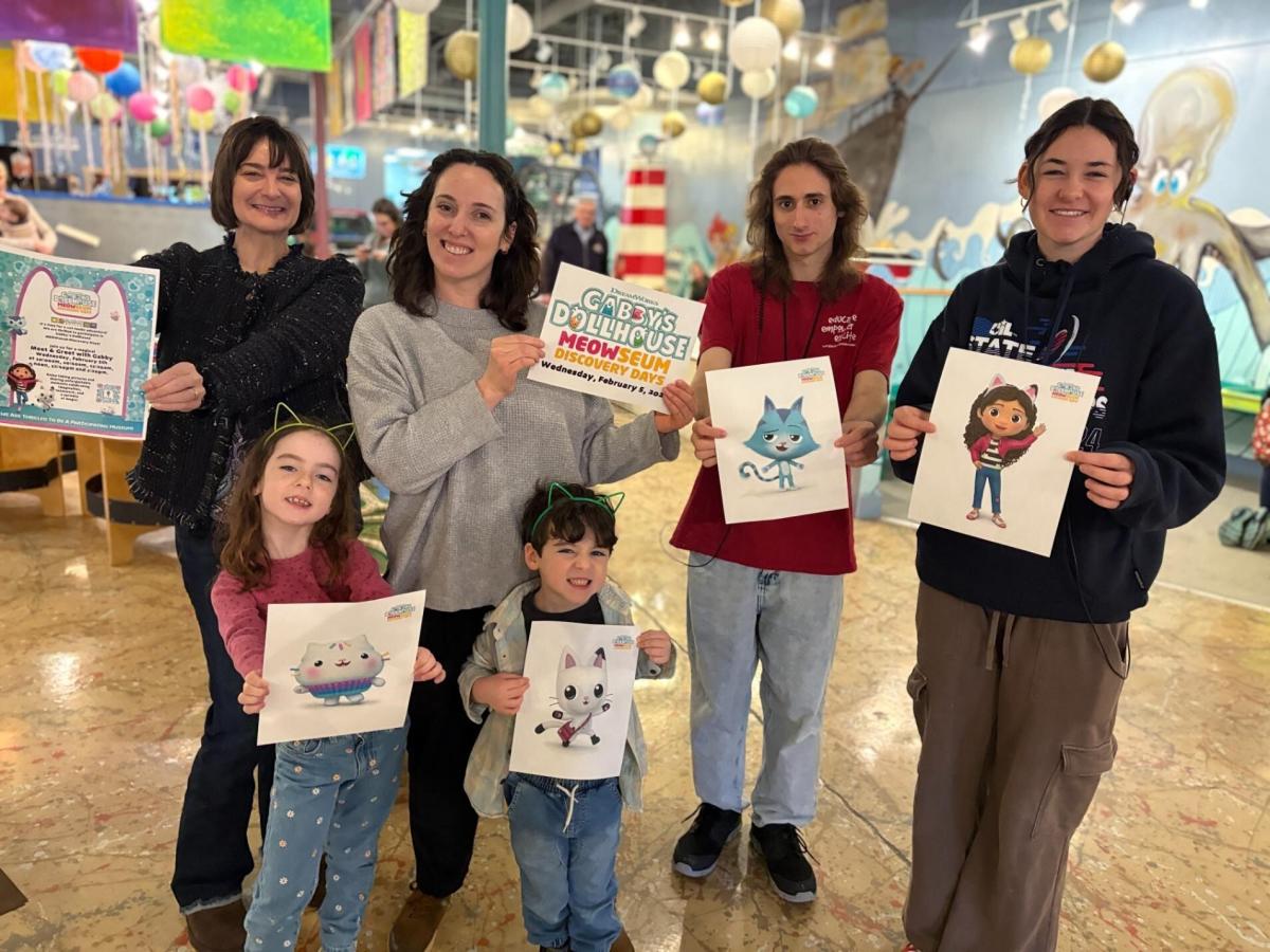 Six people in comfortable clothes (four adults in back, a young girl and boy in front) smile at the camera, each showing off a flier for the MEOWseum exhibit or a character portrait from the Gabby's Dollhouse animated TV show.