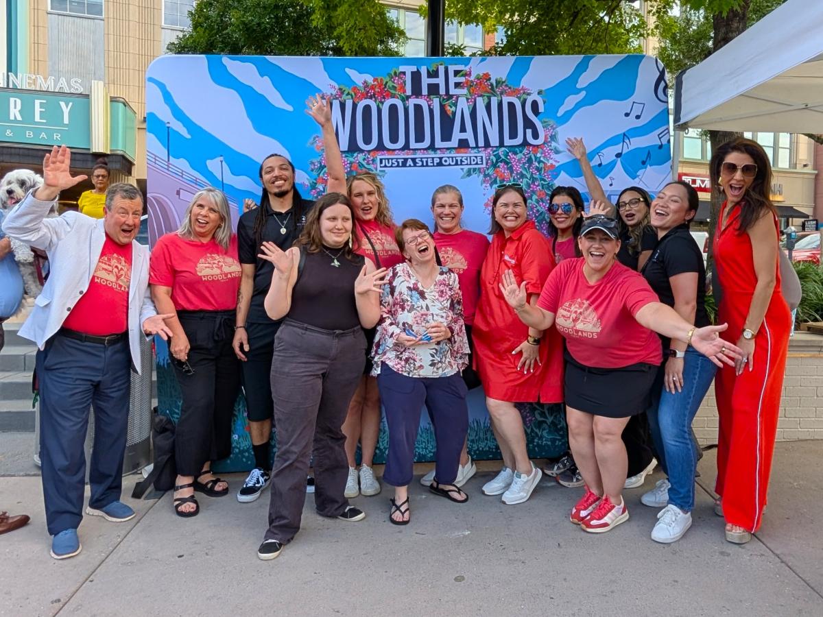 Thirteen people smile and pose for the camera in front of a backdrop of a mural of The Woodlands. Among them are Visit The Woodlands Staff Members: Nick Wolda, Julie Quinn, Josie Lewis, Ashley Fenner, Elizabeth Eddins, Carla Twidell, and Amber Parsley. Others include Market Street staff, winners of the National Travel and Tourism Week Awards, a local FOX News anchor, and more.