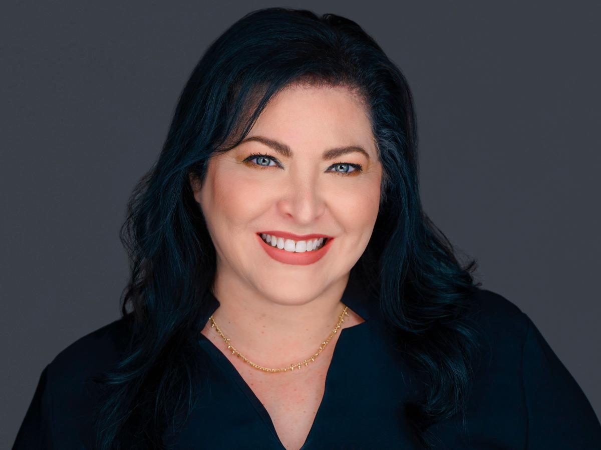 A headshot of a woman smiling at the camera. She has light blue eyes and black hair that rests on her shoulders. She is wearing a black blouse, a thin gold necklace, and natural-looking makeup. The background is solid gray.
