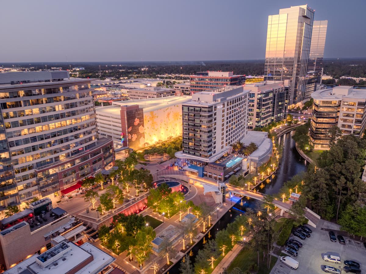 An overhead view of Waterway Square at dusk, featuring The Towers at The Woodlands Waterway, the Westin, The Fountains at The Waterway, the flower mural, a long stretch of The Waterway, and more. Most of the trees in the area are wrapped in golden lights.