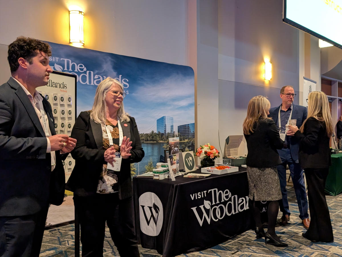 The camera focuses on the Visit The Woodlands sales booth at the 2025 Economic Outlook Conference (EOC). In the foreground are a professional businessmen Ryan Greenwood and Julie Quinn, smiling and ready to greet conferencegoers with questions about VTW. Further back are two women and a man discussing business.