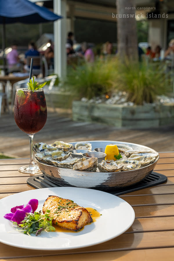 a plate of grilled fish, a dish of raw oysters, and a cocktail on an outdoor patio table