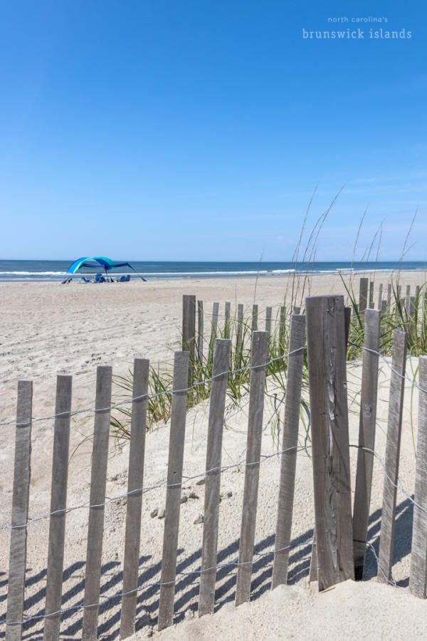 a wooden sand fence on Holden Beach, NC with a sunshade in the background