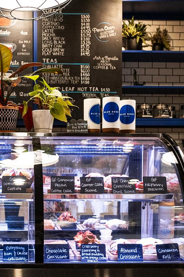 A glass display case at Blue Door Coffee Co. features a variety of seasonal dessert options, many gluten free (e.g., GF cinnamon pecan coffee cake, GF raspberry macaron, almond croissant). Atop the case are potted plants and a chalkboard featuring coffee and tea options.