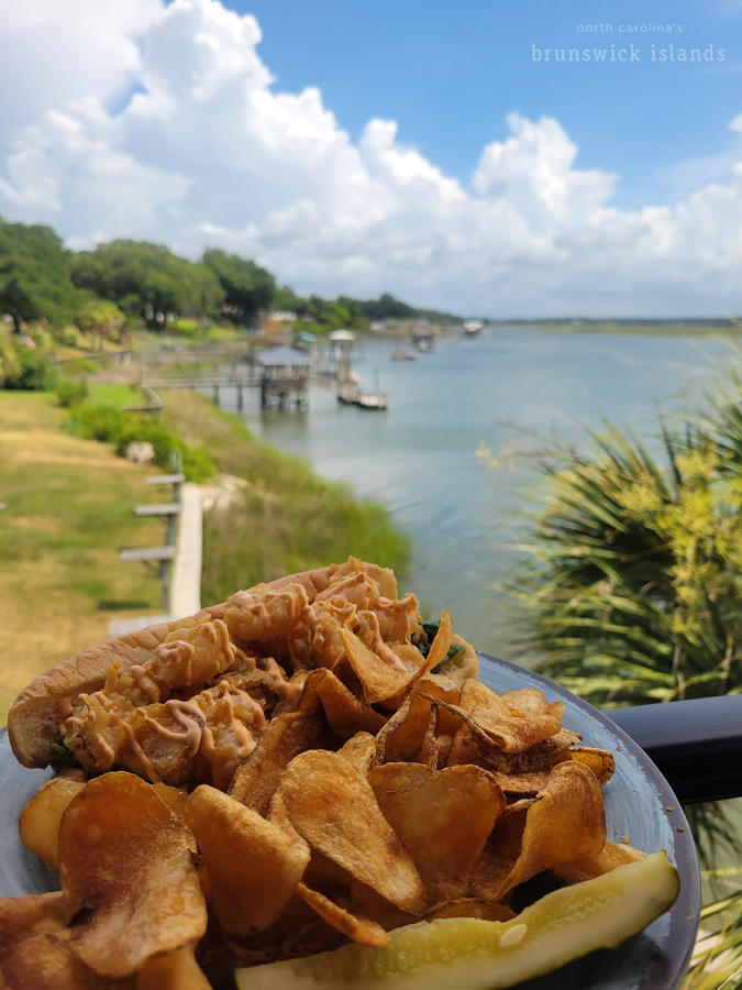 a plate with a shrimp po boy, chips, and pickle overlooking a view of the Shallotte River