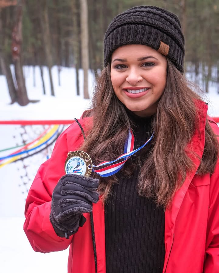 A person in a red jacket and black beanie holding a medal with a ribbon, smiling in a snowy forest setting.