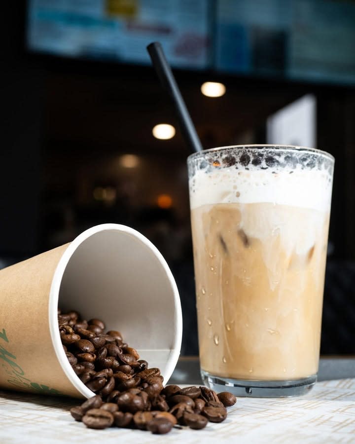 A glass of iced coffee with a straw next to a spilled cup of coffee beans, set on a table with a blurred cafe menu in the background.
